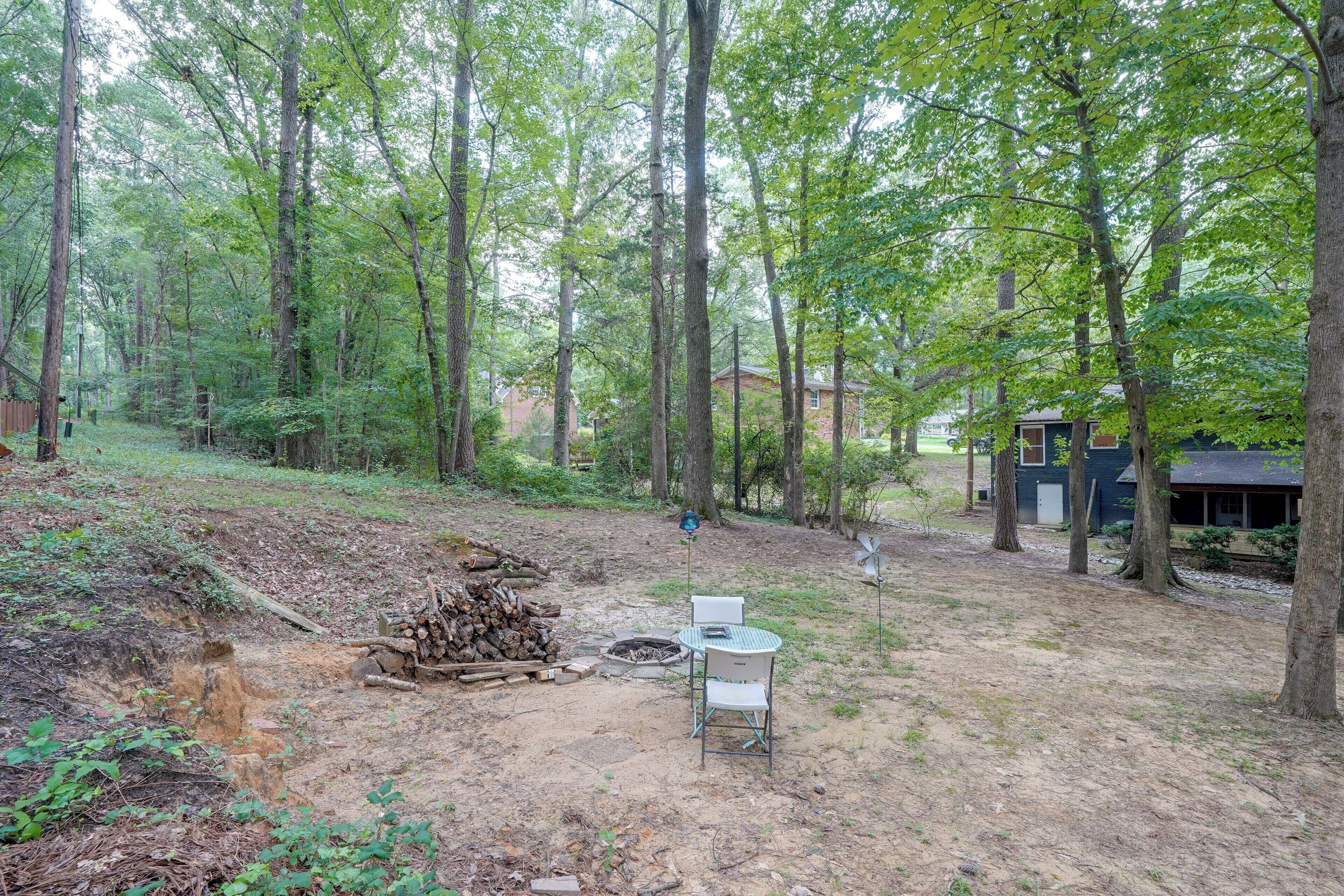 A forested backyard with a fire pit, a chair, and a stack of firewood.