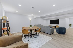 Ein modernes Wohnzimmer mit einem runden Holztisch, zwei Stühlen, einem grauen Sofa, einem blauen Hocker und einem flachbildfernseher an der Wand.