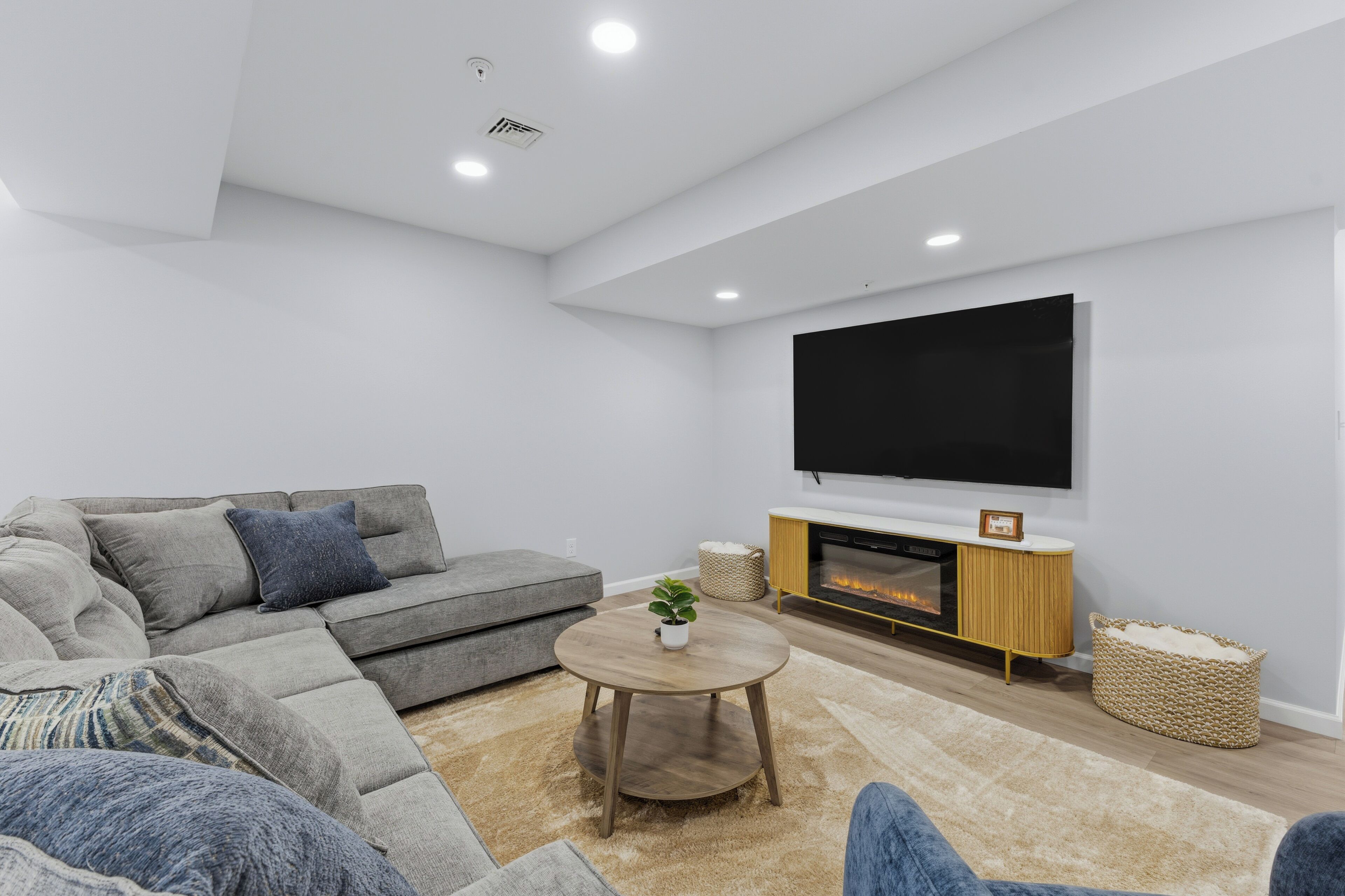 Een moderne woonkamer met een grijze modulaire bank, een ronde houten salontafel, een flatscreen televisie die aan de muur is bevestigd, en een open haard met een witte schouw.
