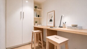 Una habitación minimalista con un escritorio de madera, dos taburetes, una estantería y un cuadro enmarcado.