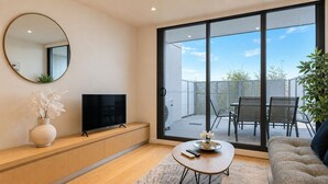 Una sala de estar moderna con un televisor de pantalla plana, un espejo redondo, un sofá y un área de comedor visible a través de una puerta de vidrio.
