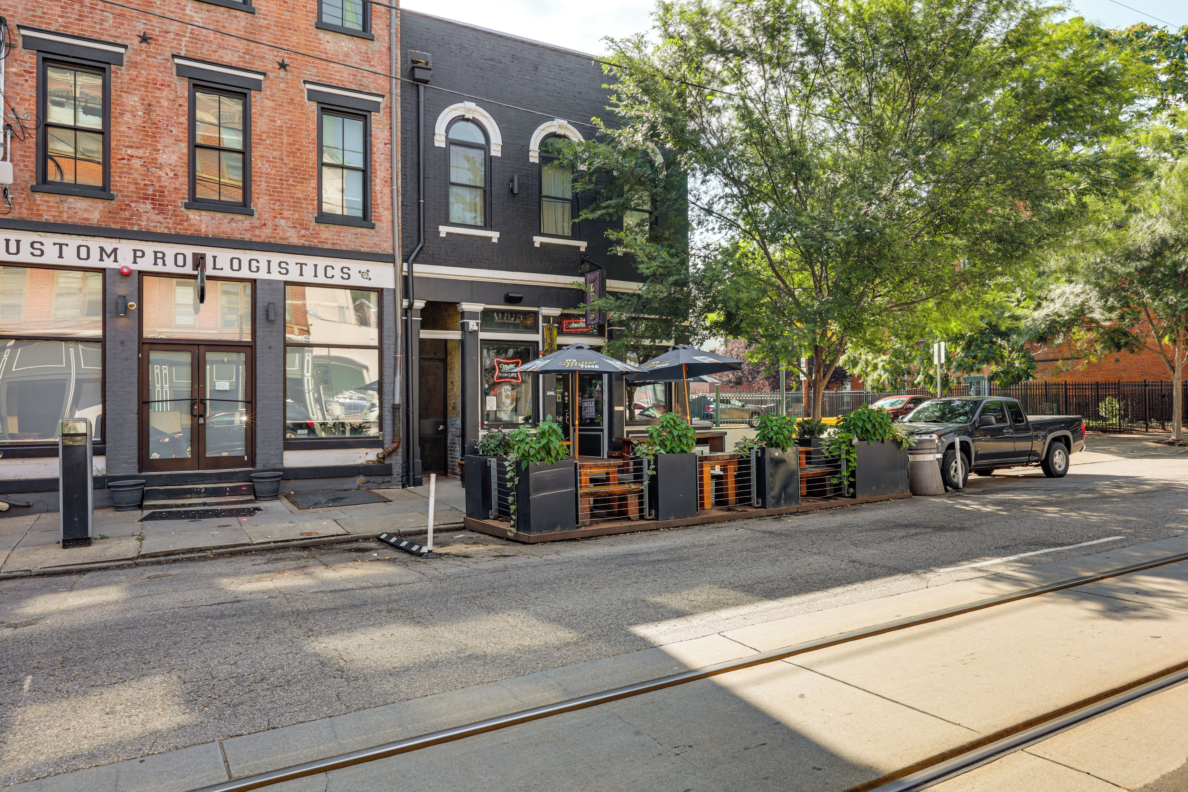 Une vue de rue avec un bâtiment portant l’inscription « CUSTOM PRO LOGISTICS » et des tables à l’extérieur, protégées par des parasols.