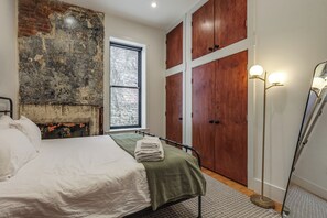 Ein Schlafzimmer mit einer Ziegelwand, einem Fenster, einem Bett mit grüner Decke, Holzschränken und einer Stehlampe.