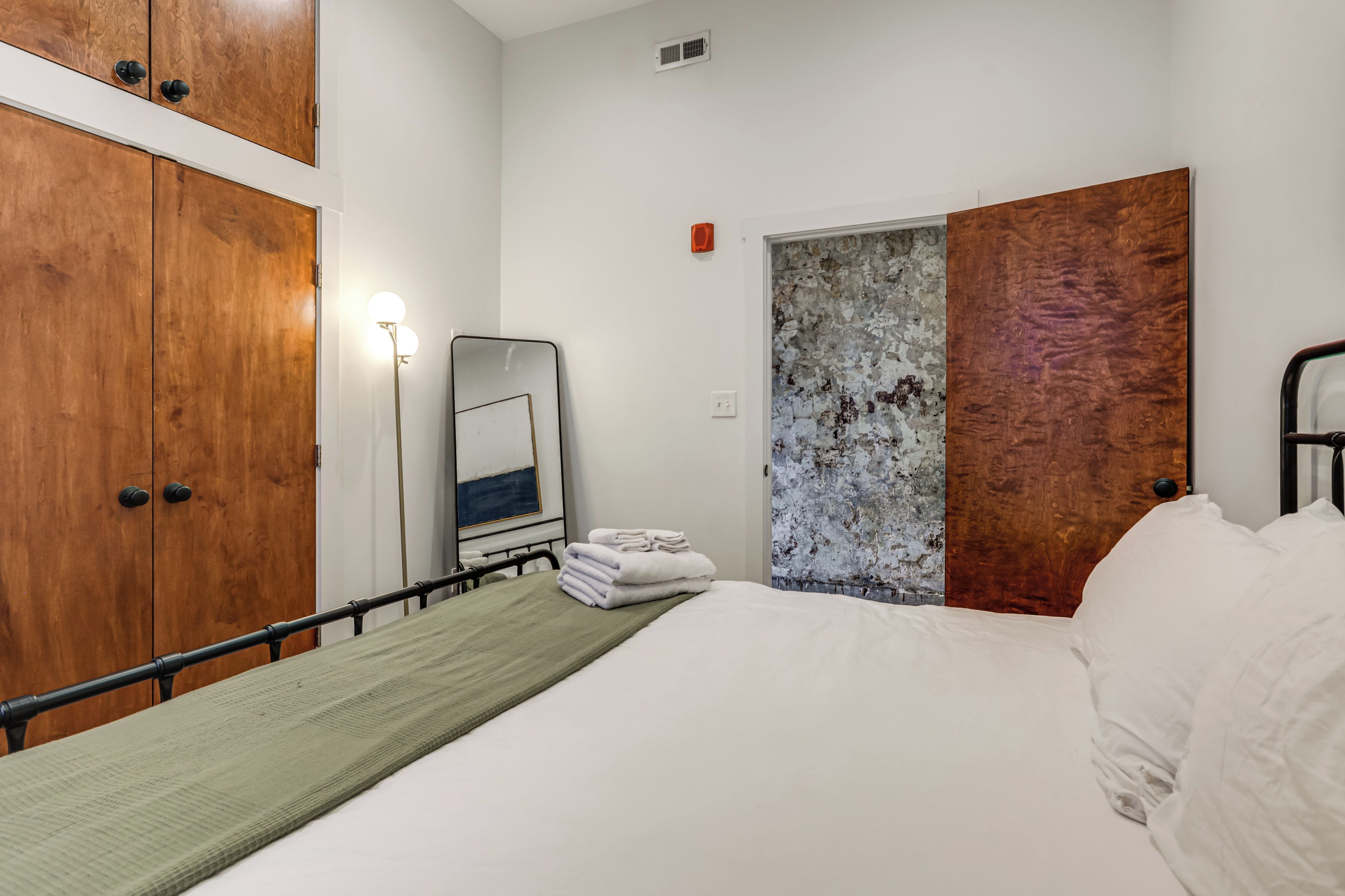 A hotel room with a bed, a bedside table, a wall-mounted mirror, a wooden wardrobe, and a door with a textured panel.