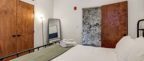 Habitación de hotel con cama, mesita de noche, espejo fijado a la pared, armario de madera y puerta con panel texturizado.