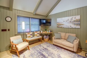 A living room with a beige sofa, a wooden chair, a bench, a flat-screen TV, a painting of ducks, and a clock on the wall. - Steps to Lake! Cabin w/ Water Views in Hot Springs (Hot Springs)
