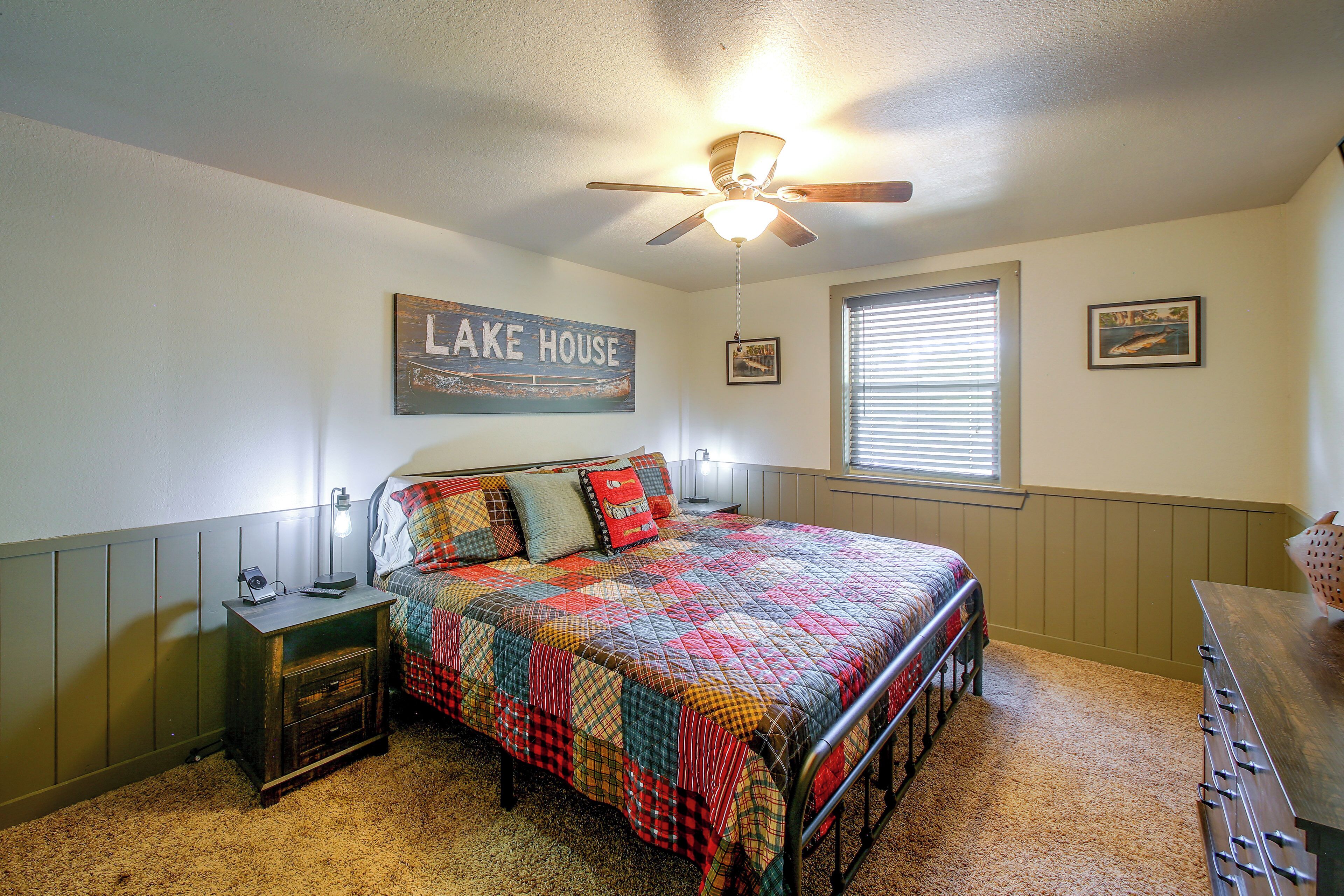 A bedroom with a bed, a nightstand, a ceiling fan, a window with blinds, and a wall with a framed picture and a sign.
