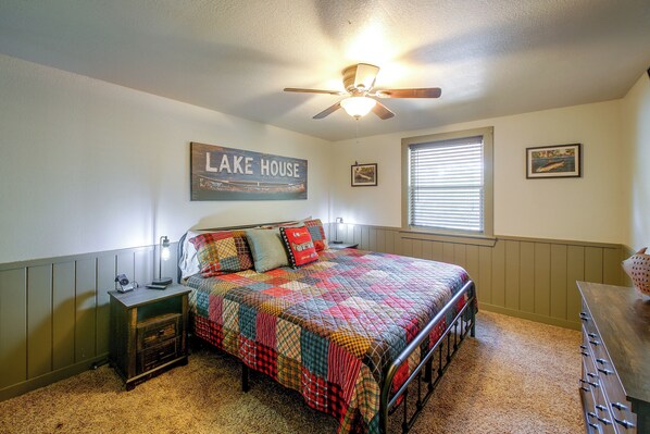 A bedroom with a bed, a nightstand, a ceiling fan, a window with blinds, and a wall with a framed picture and a sign. - Steps to Lake! Cabin w/ Water Views in Hot Springs (Hot Springs)