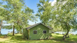 A small green cabin with a sloped roof, surrounded by lush trees and a body of water. - Pet-friendly Rustic Cabin Steps From Lake Chetek (Chetek)