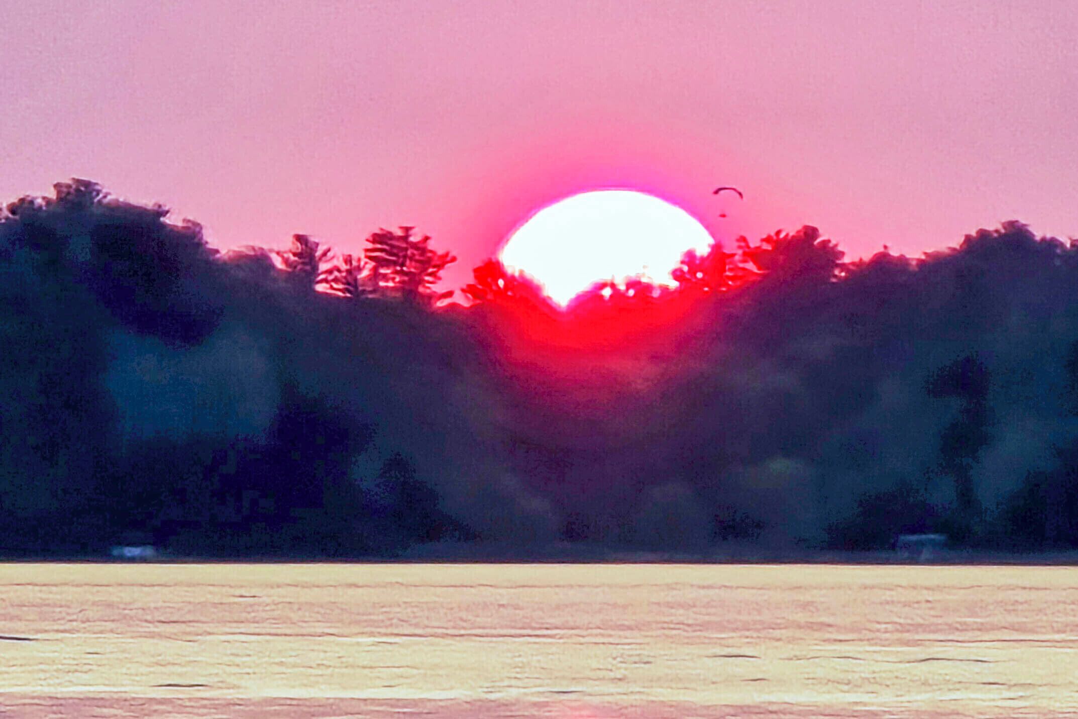 Ein Sonnenuntergang über einem Gewässer, im Vordergrund Bäume.
