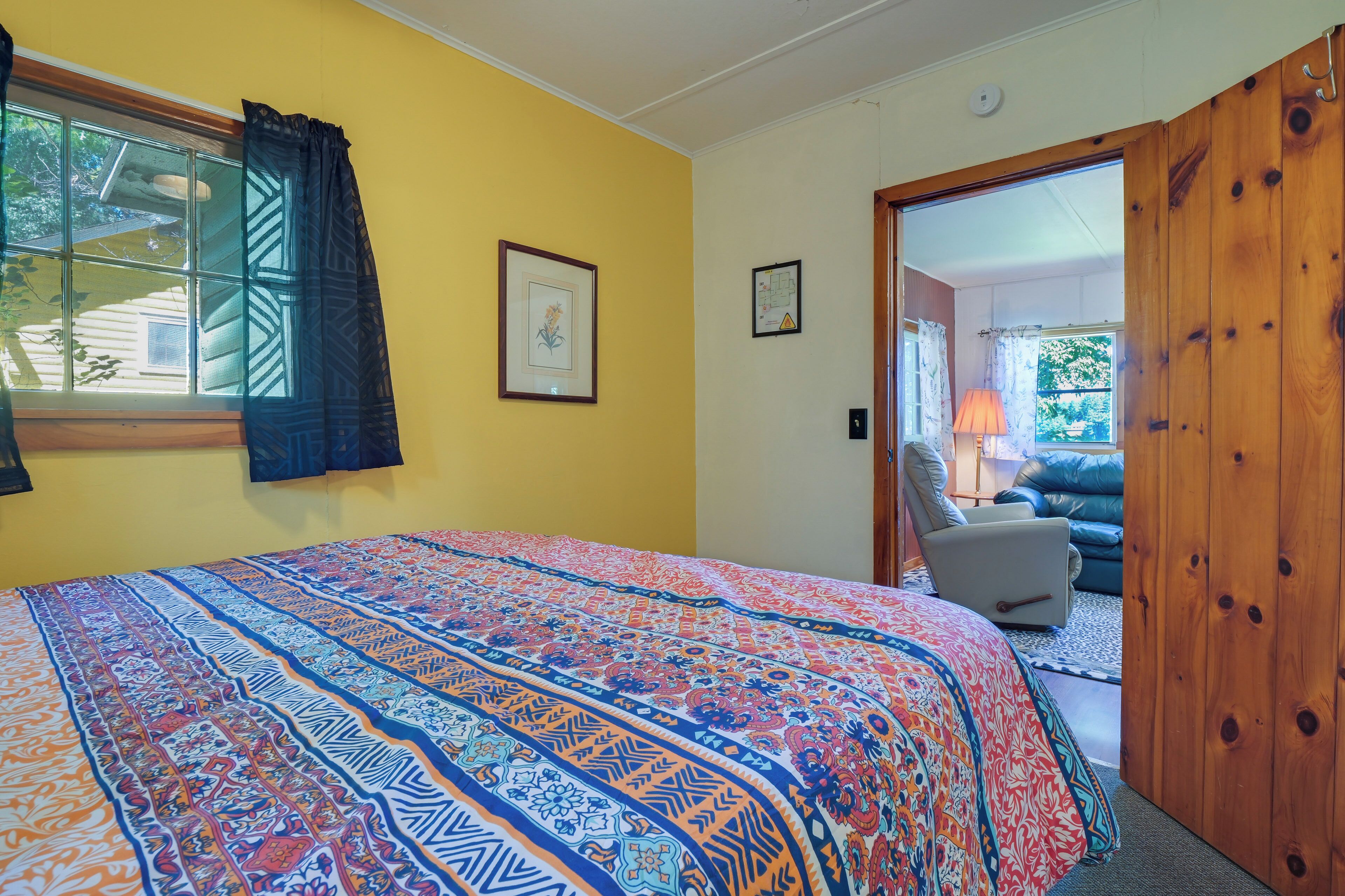 A bedroom with a bed, a window with curtains, a door leading to another room, and a wall-mounted light switch.