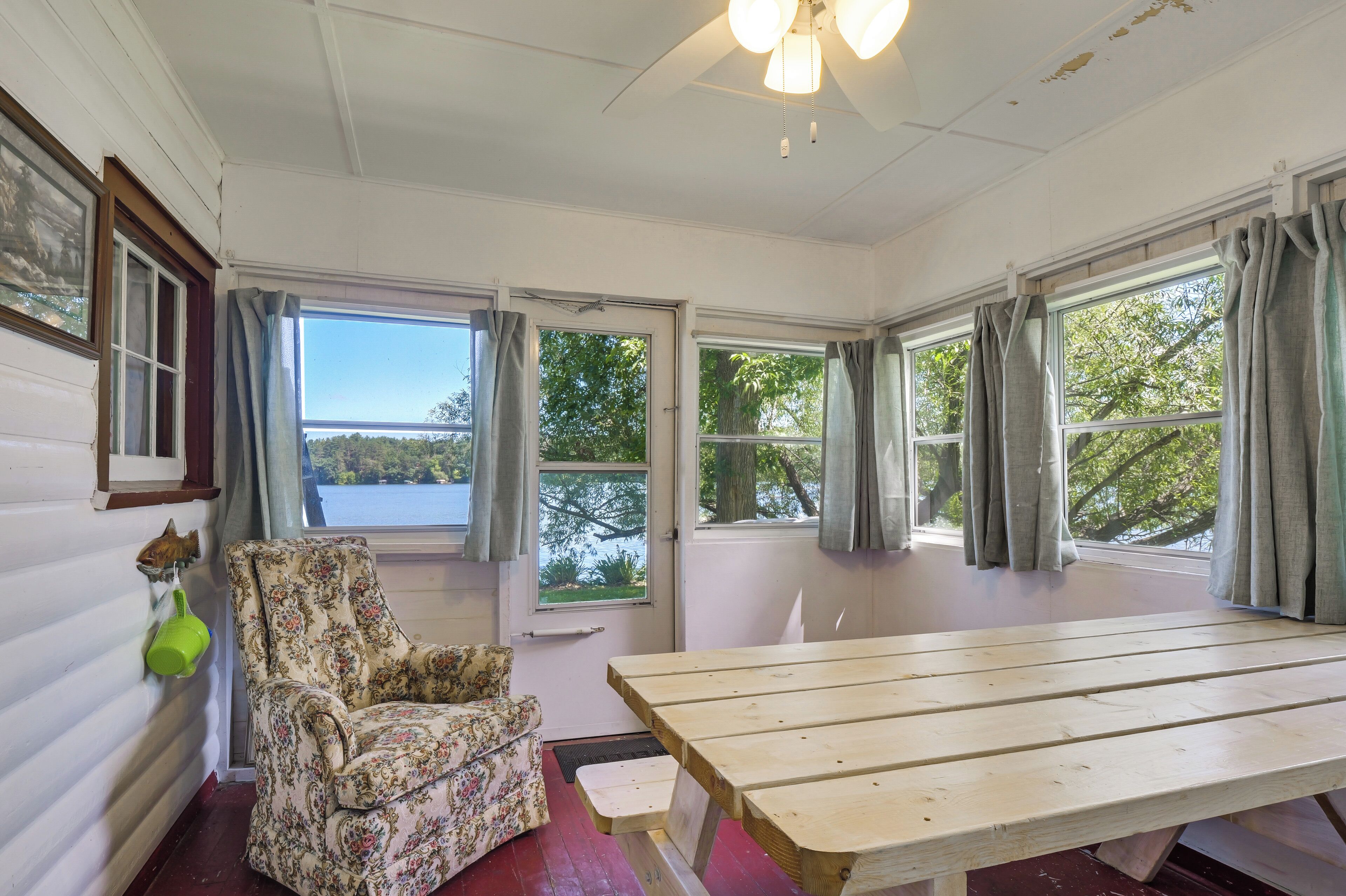 A room with a wooden table, a floral armchair, and a view of a lake through the windows.