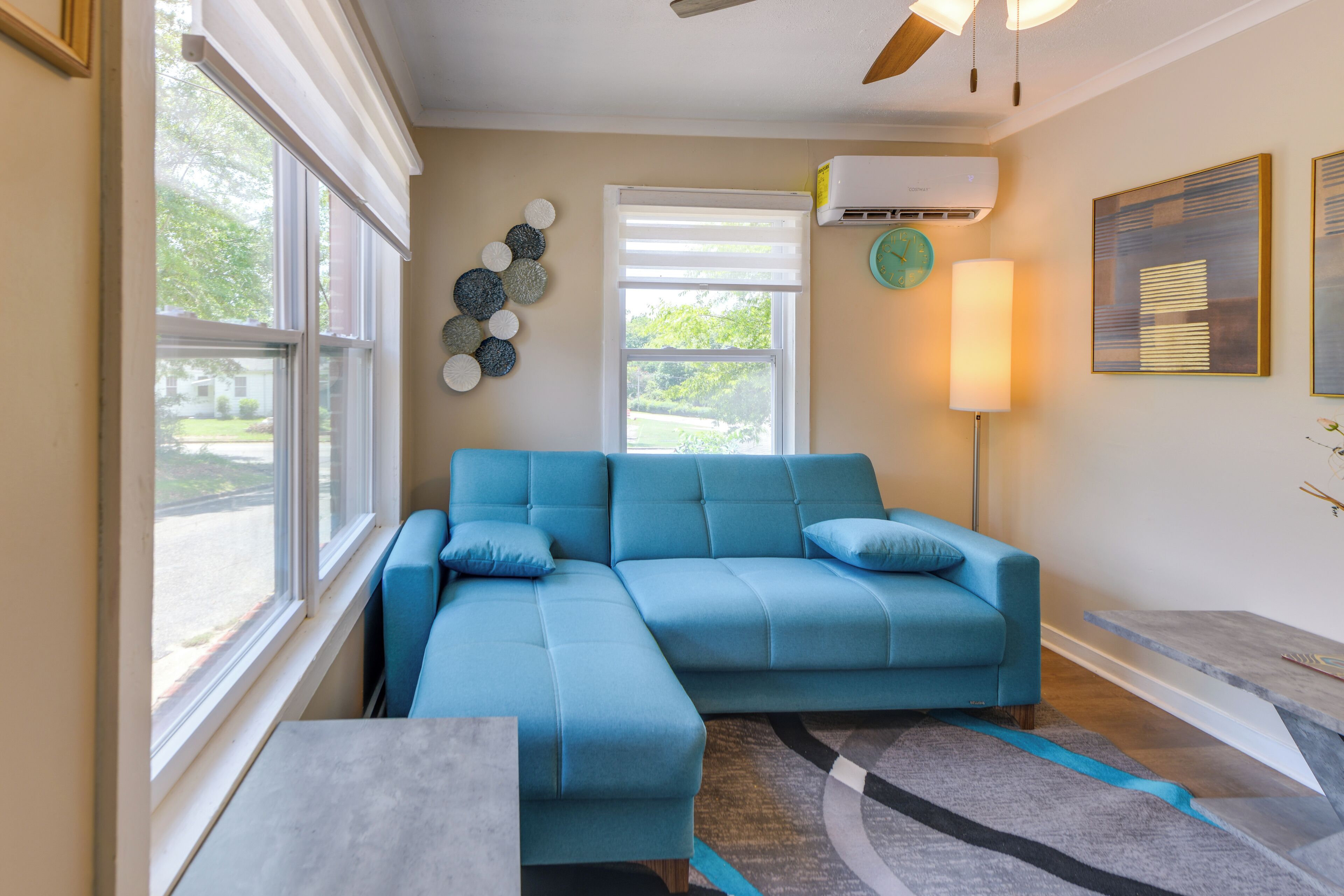 A living room with a blue sectional sofa, a side table, a lamp, a clock, and an air conditioning unit.