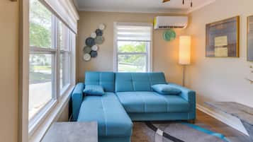 A living room with a blue sectional sofa, a side table, a lamp, a clock, and an air conditioning unit.