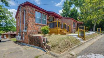 A two-story brick house with a red roof, a front porch with a wooden staircase, and a small front yard.