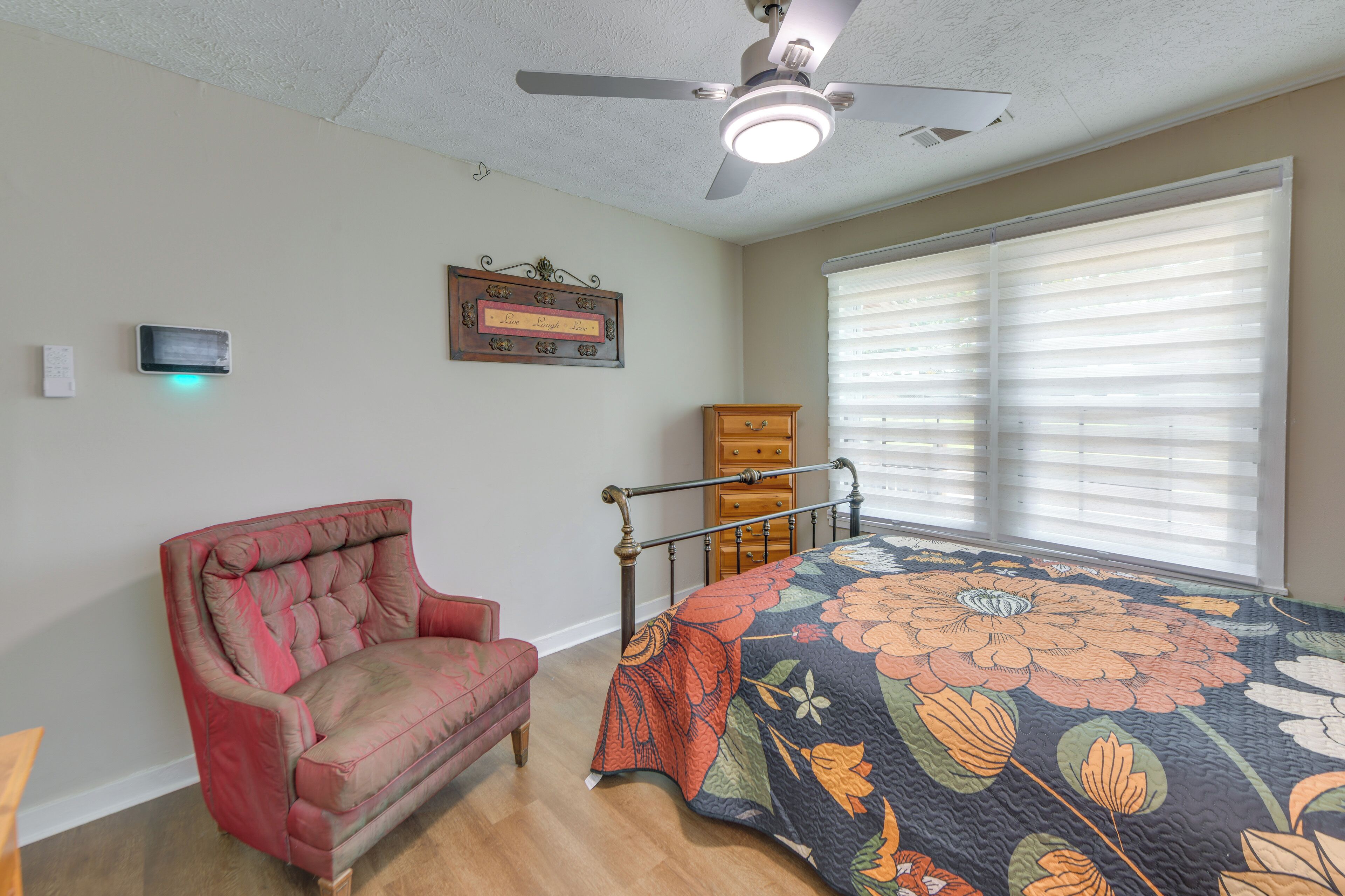 A bedroom with a bed, a chair, a ceiling fan, a window with blinds, and a wall-mounted thermostat.