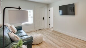 A modern living room with a leather sofa, a floor lamp, a flat-screen TV mounted on the wall, and a wooden floor. - Mtn-view Deck w/ Grill! Modern Medicine Park Cabin (Medicine Park)