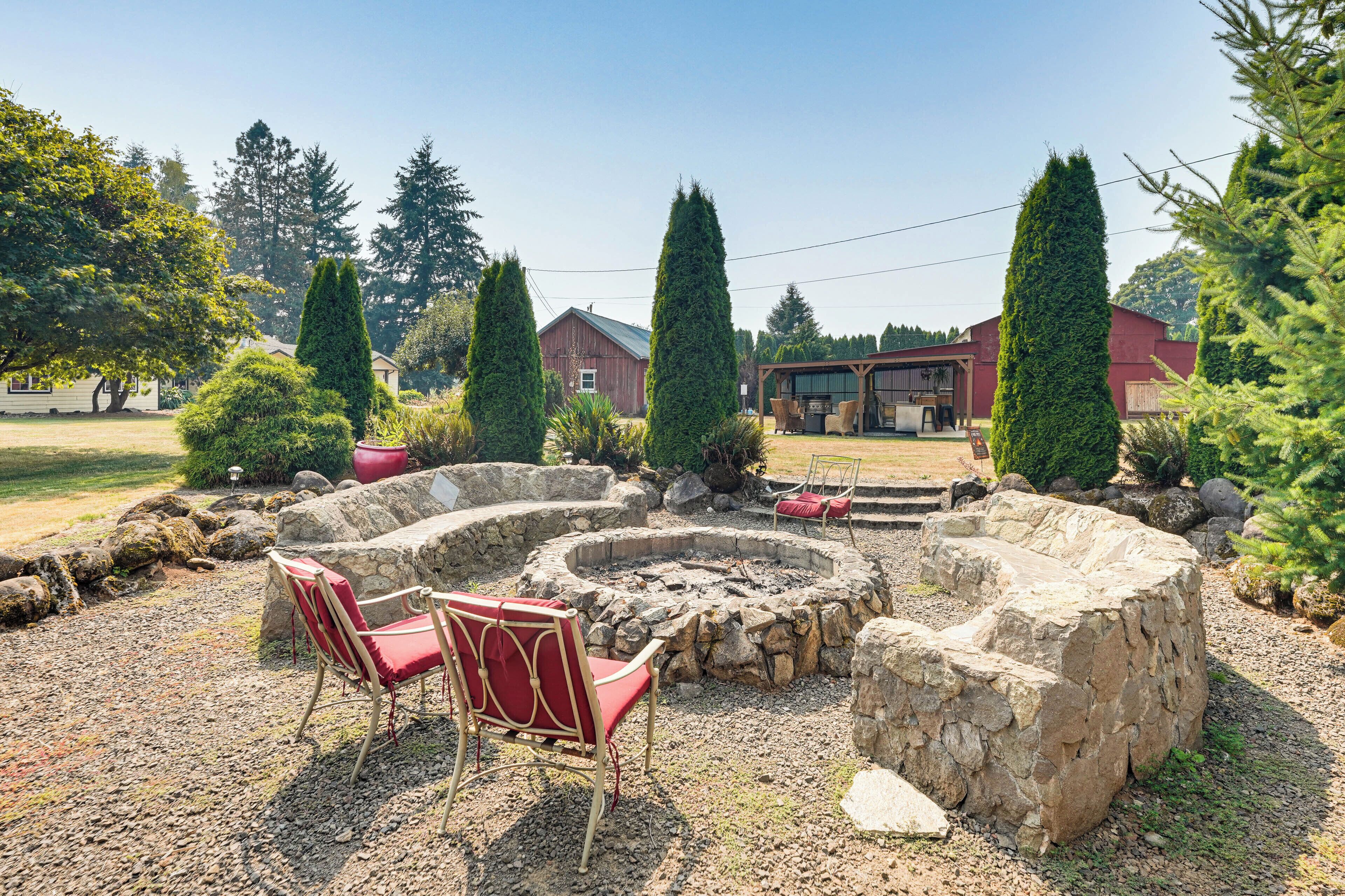 A stone fire pit with two red chairs, surrounded by rocks and greenery, with houses in the background.
