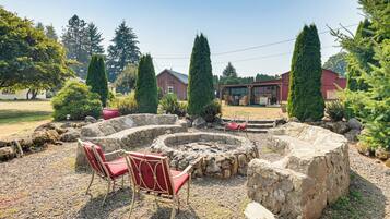 A stone fire pit with two red chairs, surrounded by rocks and greenery, with houses in the background.