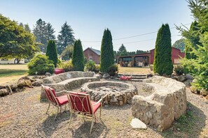 A stone fire pit with two red chairs, surrounded by rocks and greenery, with houses in the background.