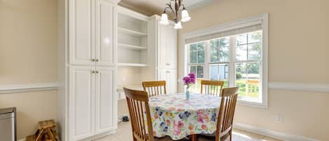A dining area with a table set for four, surrounded by wooden chairs, a floral tablecloth, and a vase with flowers.