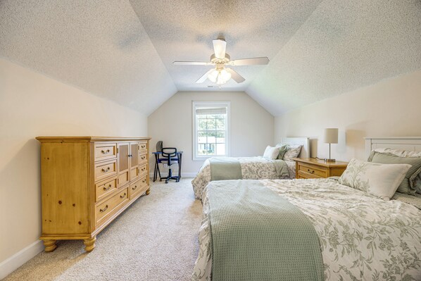 A bedroom with a sloped ceiling, a ceiling fan, a desk, two beds, a dresser, and a window.