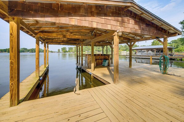 Un muelle de madera que se adentra en un lago tranquilo, con un espacio de estar cubierto y un bote amarrado a la derecha.