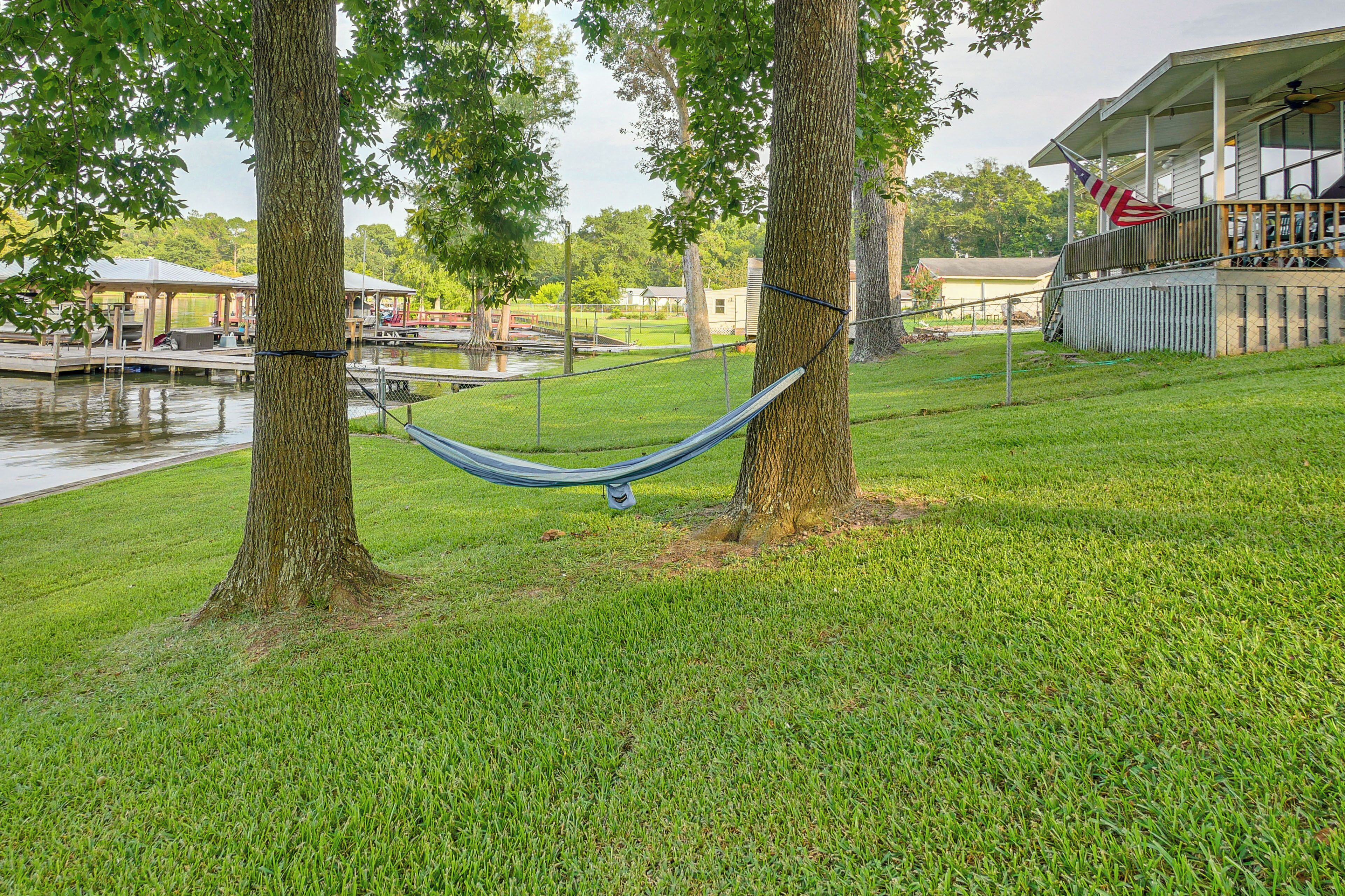 A hammock between two trees in a grassy area with a dock and a building in the background.