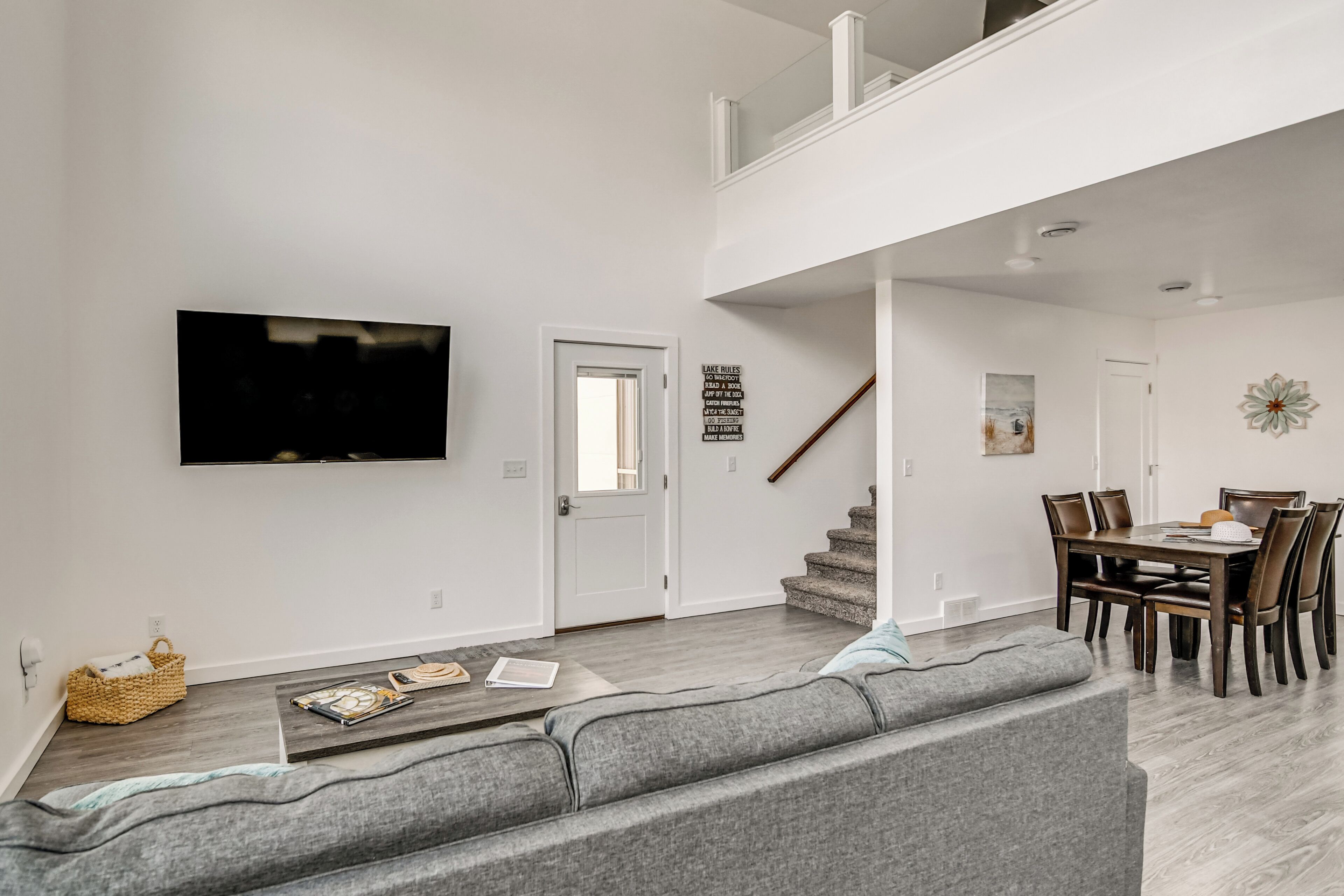 Una sala de estar moderna con un sofá gris, un televisor de pantalla plana montado en la pared, una zona de comedor con una mesa y sillas de madera, y una escalera que lleva a un nivel superior.