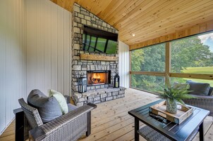 A stone fireplace with a flat-screen TV above it, a wicker sofa with cushions, a wooden table with a tray, and a large window with a view of trees.