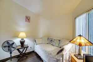 A bedroom with a bed, a bedside table with a lamp, a fan, and a window with blinds.