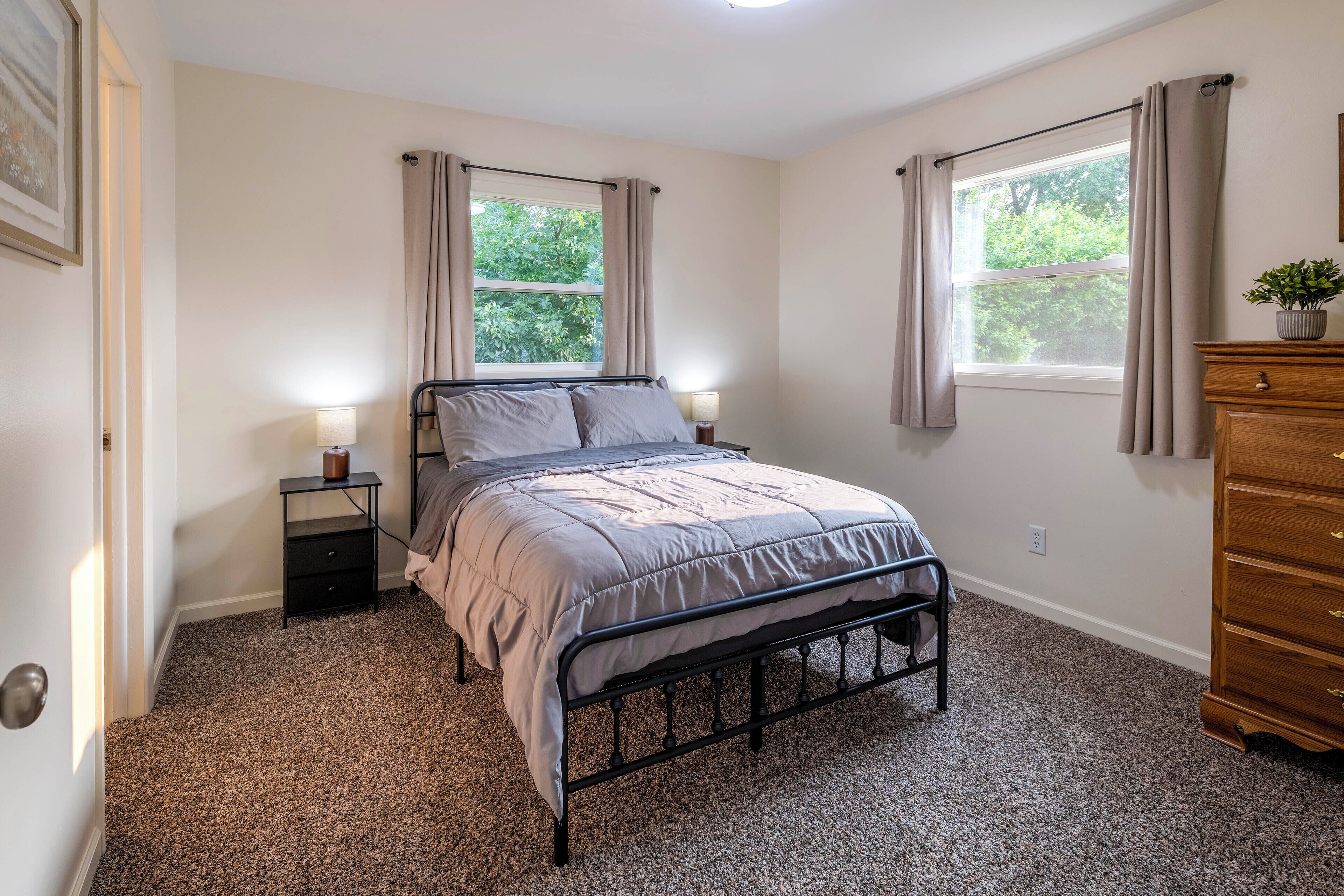 A bedroom with a bed, bedside tables, a dresser, and two windows with curtains.