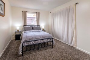 A bedroom with a bed, two bedside tables, a window with curtains, and a carpeted floor.