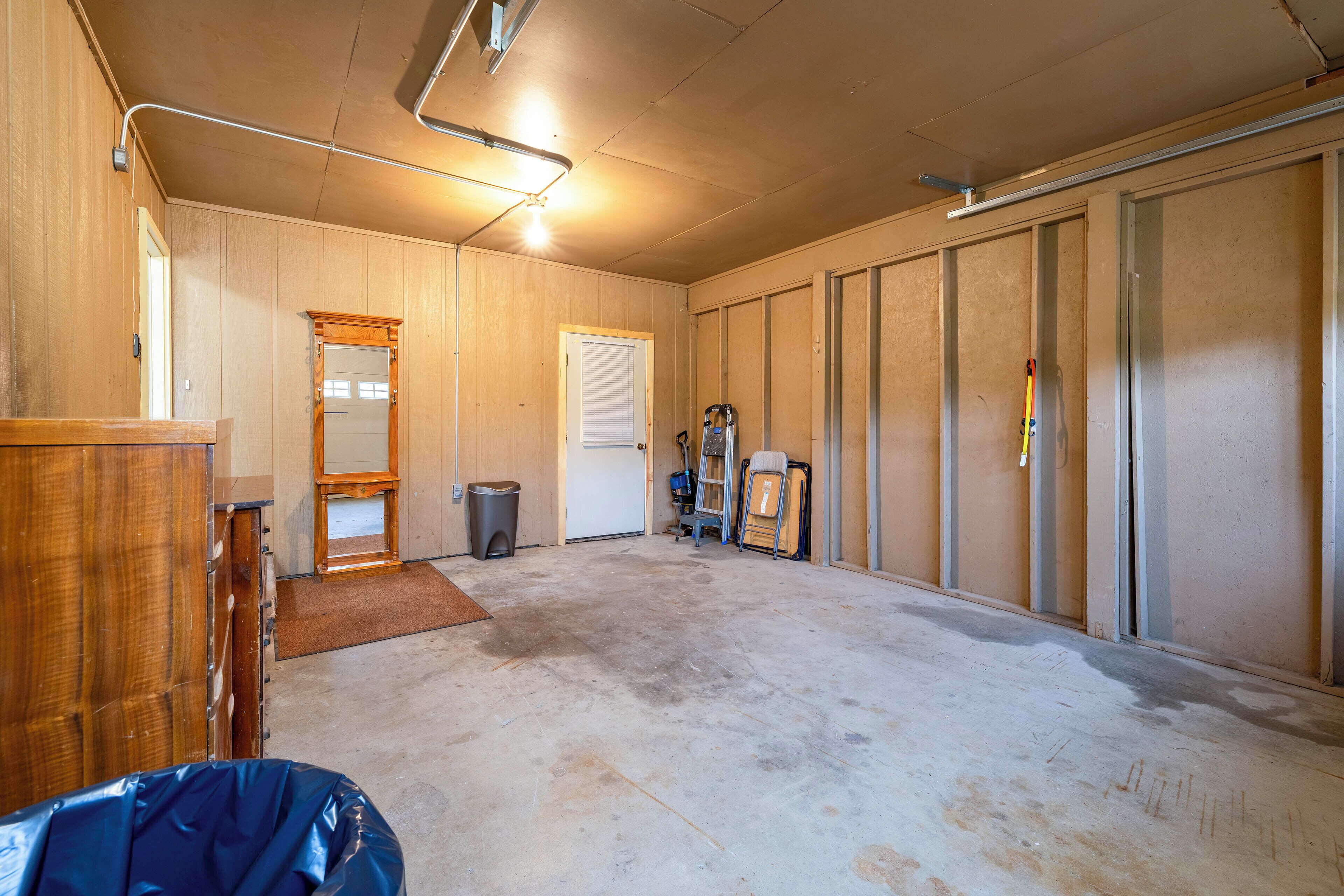 A spacious garage with a concrete floor, wooden walls, a large mirror, a door, and various tools and equipment.