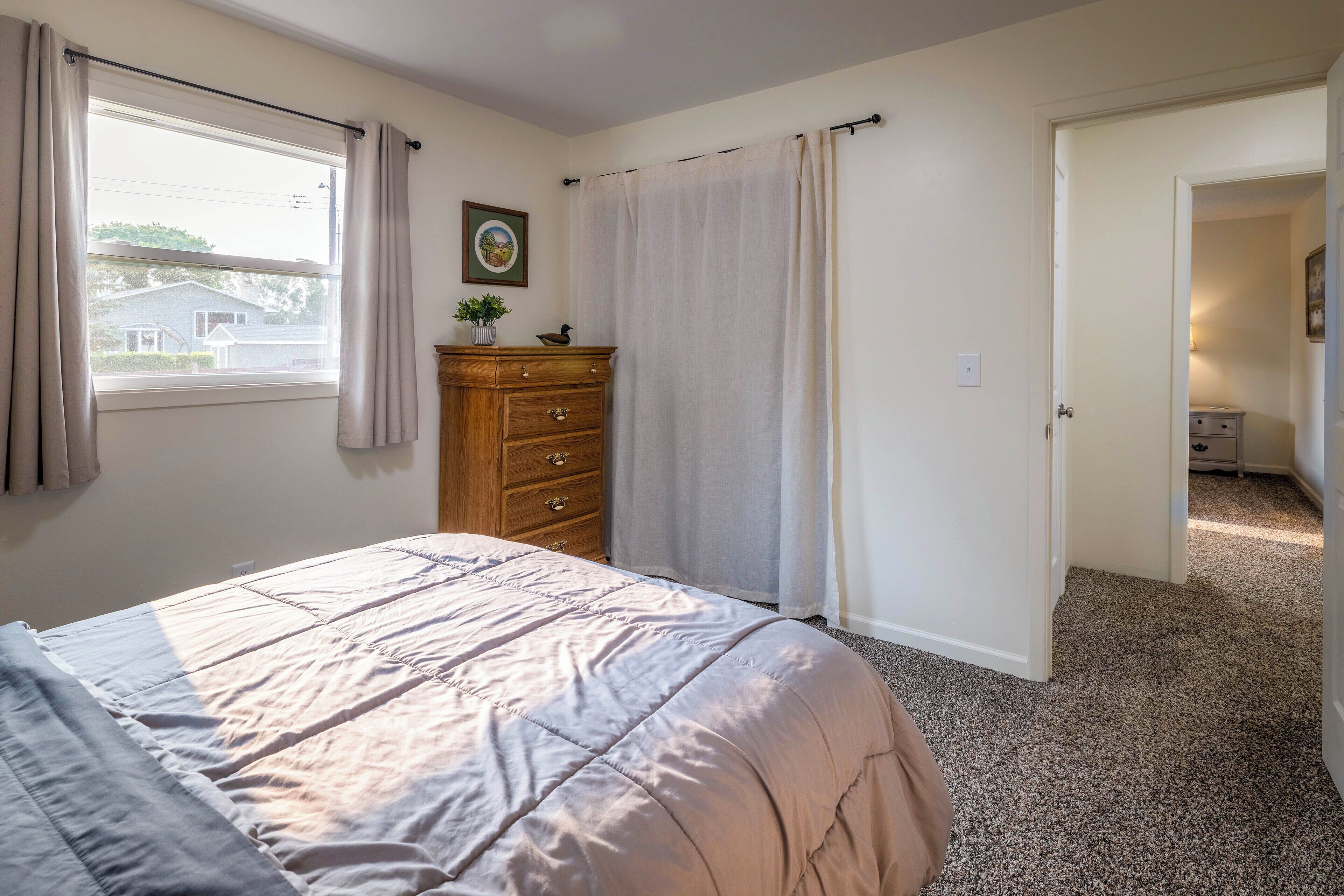 A bedroom with a bed, a dresser, a window with curtains, a door leading to another room, and a framed picture on the wall.