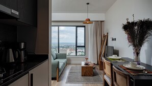 Eine moderne Küche mit Kaffeemaschine, ein Essbereich mit Tisch für zwei Personen, eine Sitzecke und Blick auf die Stadt durch große Fenster.
