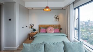 A modern bedroom with a large bed, a nightstand, a lamp, and a view of the city through a large window. - Stylish Studio apartment Upperhill｜Pool & Gym (Nairobi)