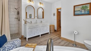 Un baño con ducha, un tocador blanco con dos espejos y un cuadro en la pared.