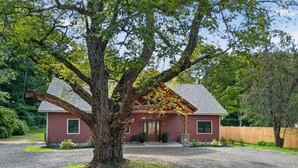 Una casa roja con un gran árbol en primer plano, un camino de grava y una valla de madera al fondo.