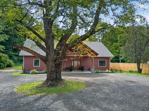 Una casa roja con un gran árbol en primer plano, un camino de grava y una valla de madera al fondo.
