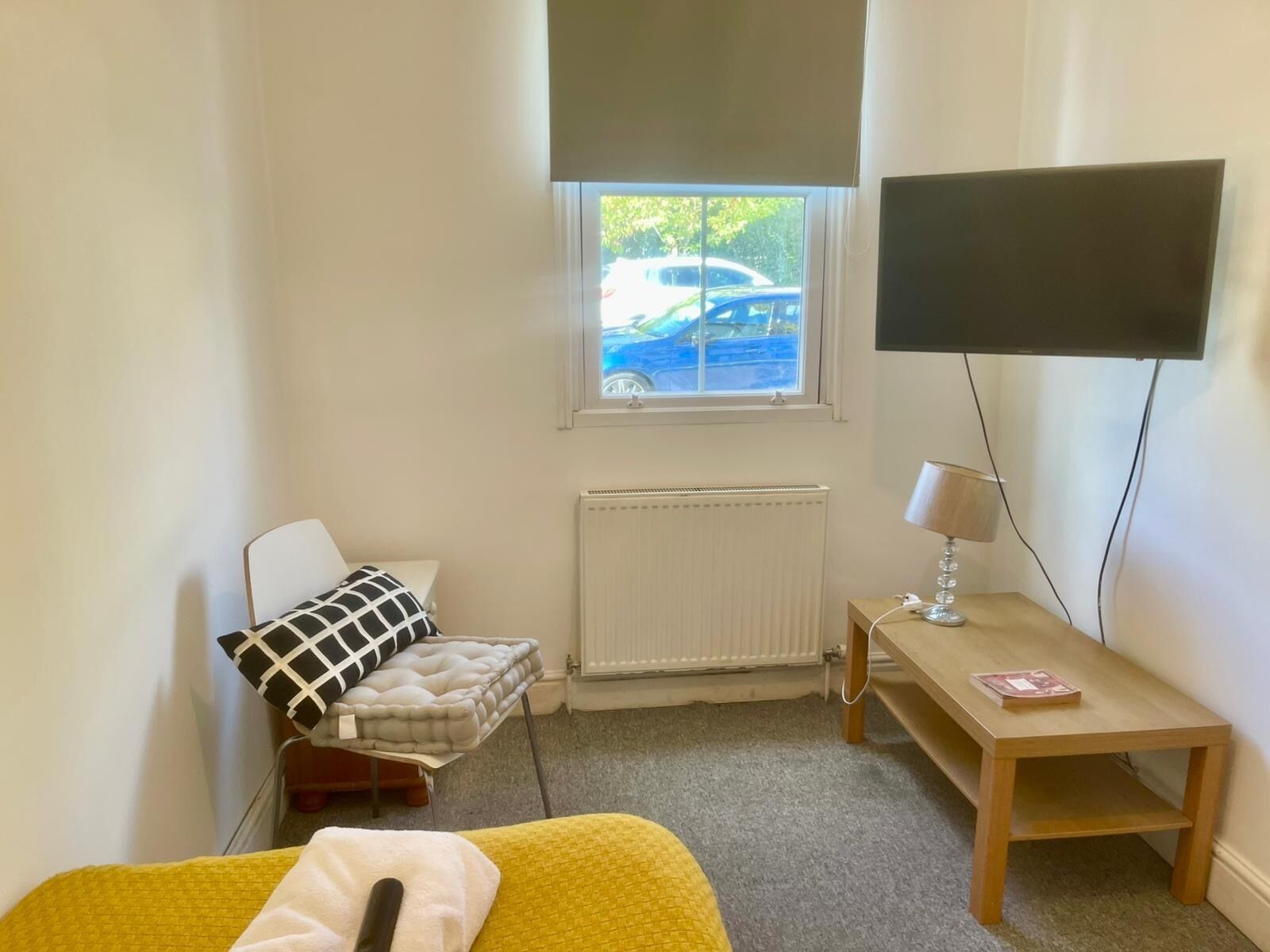 A bedroom with a bed, a bedside table with a lamp, a radiator, and a window with a view of a car outside.