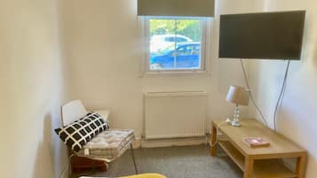 A bedroom with a bed, a bedside table with a lamp, a radiator, and a window with a view of a car outside.