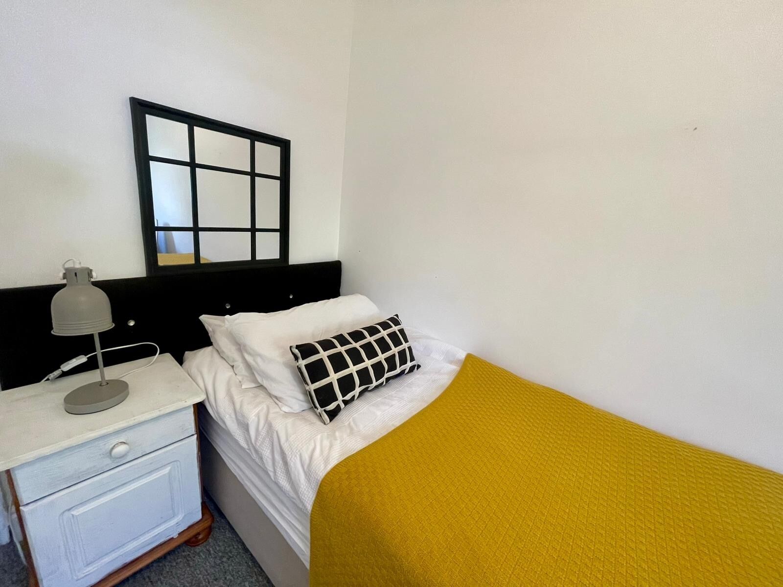 A single bed with a white mattress, a black and white pillow, and a mustard-colored blanket. There is a bedside table with a lamp and a small drawer. A black-framed mirror hangs on the wall above the bed.