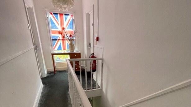 A staircase with a white handrail leading to a landing with a vase of flowers and a window displaying a flag.