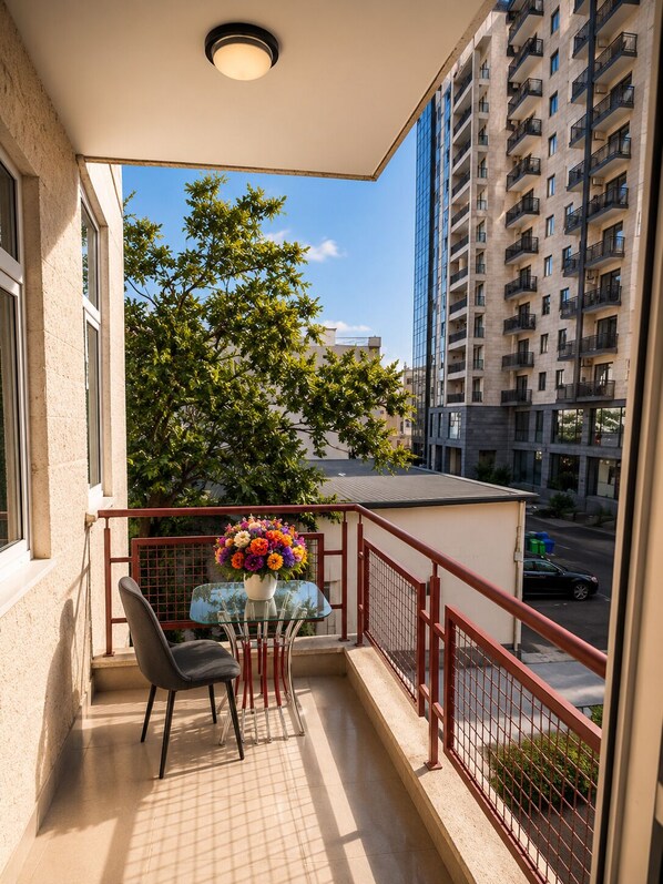 A balcony with a table and chairs, a flower arrangement, and a view of a tall building and tree.