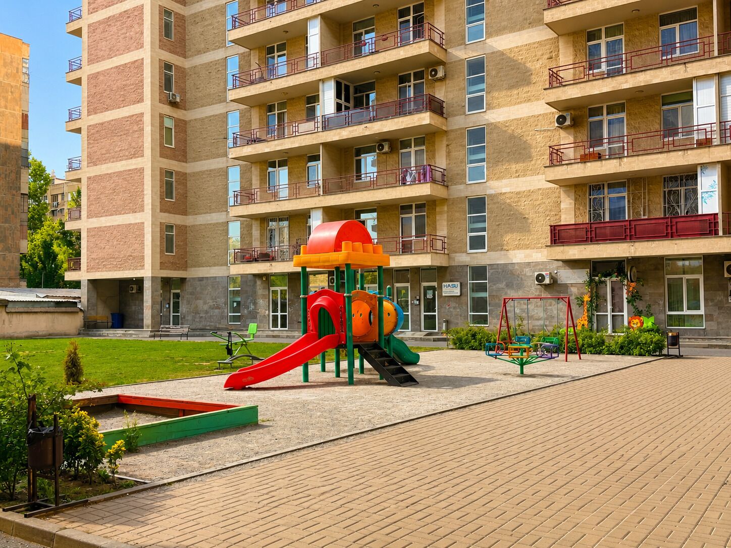 A multi-story residential building with balconies, a playground with a slide, and a paved area with benches.