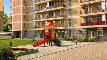 A multi-story residential building with balconies, a playground with a slide, and a paved area with benches.