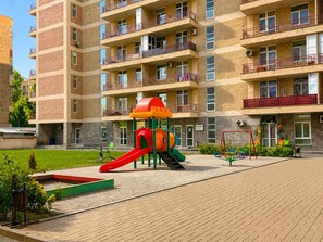 Ein mehrstöckiges Wohnhaus mit Balkonen, ein Spielplatz mit Rutsche und ein gepflasterter Bereich mit Bänken.