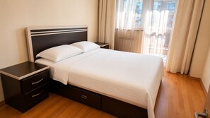 A neatly made bed with a headboard, a bedside table with drawers, and a window with curtains.