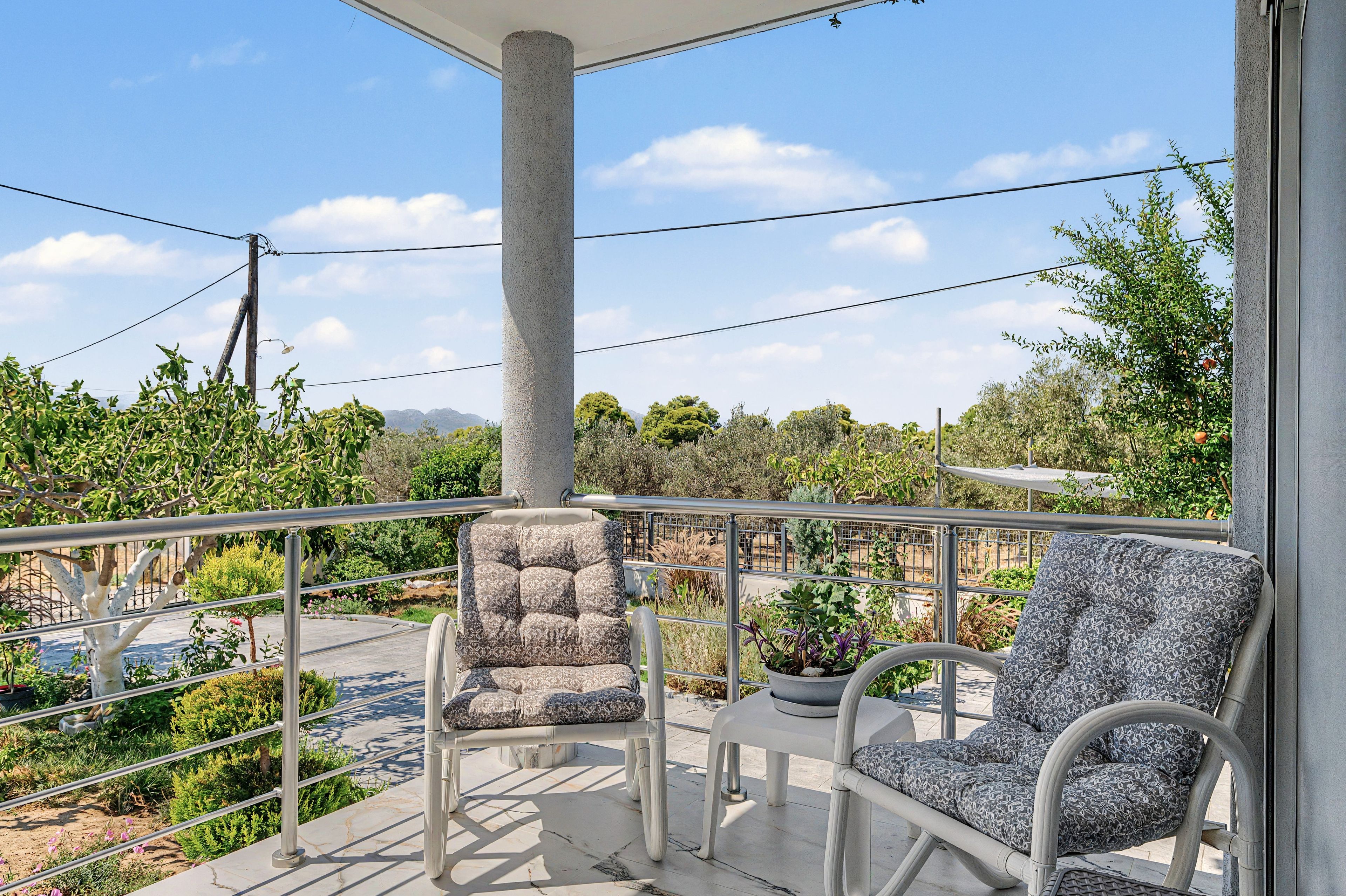 Un balcon avec deux chaises et une table, donnant sur un jardin où poussent différentes espèces de plantes et sur une chaîne de montagnes lointaine.
